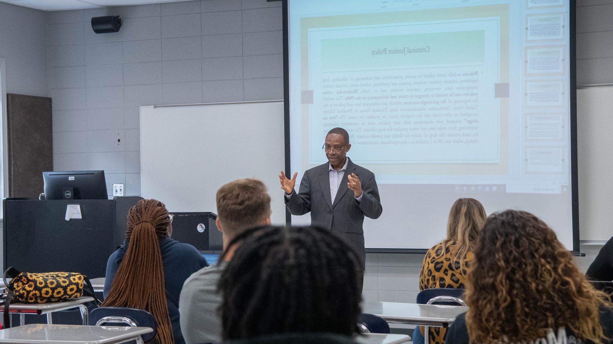 Professor giving lecture to class with PowerPoint projected behind him.